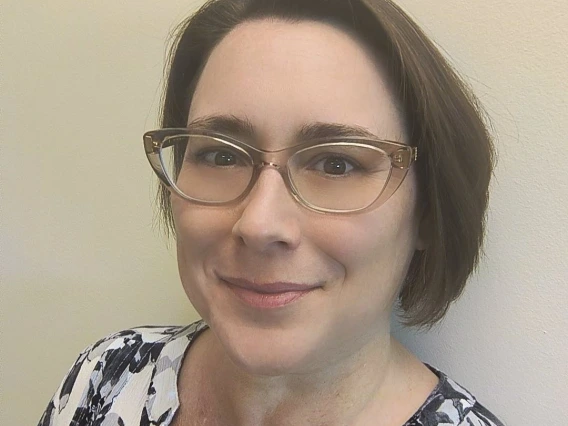 A headshot of a caucasian woman with pale skin, a straight brunette bob, and glasses wearing a black and white flowered shirt