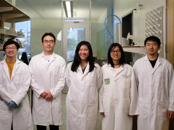 Group of five people wearing white lab coats standing in a laboratory setting. They are positioned in a line and smiling, with shelves of lab equipment visible on one side and a glass door in the background. The individuals appear to be part of a research team.