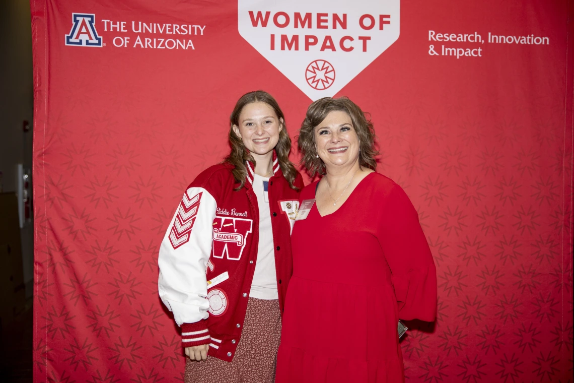 Two women pose for a photo in front of a red backdrop.