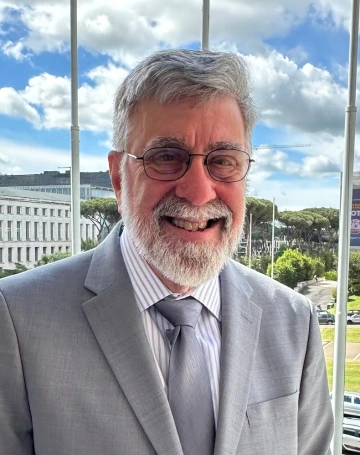 man wearing a gray suit, smiling, standing in front of white clouds and blue sky