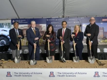 A group of six people stands under a tent, each holding a ceremonial shovel in preparation for a groundbreaking event. They are standing in front of a banner with the University of Arizona Health Sciences logo and a rendering of a new facility. Each person is dressed in business attire, and they smile as they pose for the photo. Helmets are lined up on the stage behind them.